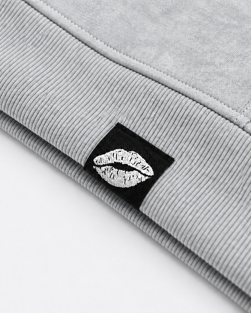 Close-up of a folded light gray ribbed sweatshirt or hoodie showing a black square patch with a white lips logo and the text 'M.G Fice O'N' on it, set against a plain white background.